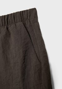 Section de pantalon en tissu de lin marron foncé avec taille élastique et coutures visibles des poches latérales sur un fond clair.