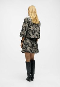 Kvinde med langt blondt hår iført en sort og guld blomstret jakke og nederdel, sorte højhælede knæstøvler og et sort armbånd, stående.