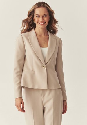 Ženska z valovitimi lasmi nosi bež prilagojeno srajco in ujemajoče hlače, pod njimi belo majico, nasmejana na enostavni svetli podlagi.