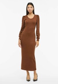 Ruskea neulemekko, jossa pitkät pussihihat, pyöreä pääntie ja vartalonmyötäinen siluetti, ulottuen puoliväliin pohjetta. Päällä mustat terävät korkokengät.