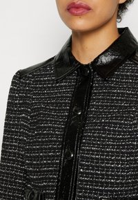 Buste supérieur d’une femme portant une veste texturée noire et blanche avec un col en cuir noir brillant, des accents aux épaules et une patte de boutonnage.