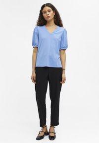 Helles blaues V-Ausschnitt-Oberteil mit gepufften kurzen Ärmeln, kombiniert mit schwarzen schmalen Hosen und schwarzen Schuhen mit Riemen. Glattes Material, simples Design.