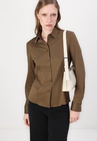 Brown long-sleeve button-up shirt with a sleek fit, featuring a pointed collar and contrasting white stitching, paired with a light beige handbag.