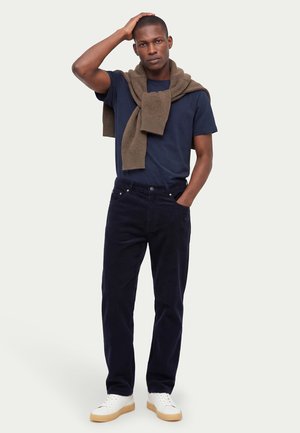 Homme portant un t-shirt bleu marine, un pantalon foncé, des baskets blanches, avec un pull marron posé sur les épaules, debout avec une main sur la tête.