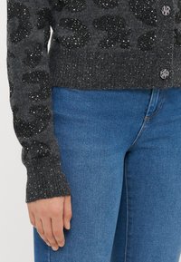 Cardigan grigio a fantasia leopardata con paillettes metalliche, polsini a coste e chiusura frontale con bottoni. Abbinato a jeans blu di denim.