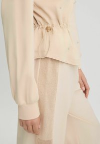Haut beige avec taille élastique, manches longues. Présente un panneau en tricot texturé sur le pantalon et des coutures décoratives. Apparence de tissu lisse.