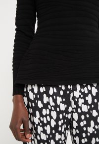 Schwarzes langärmliges Oberteil mit texturierten horizontalen Rillen, kombiniert mit schwarz-weißen gepunkteten Hosen. Nahaufnahme der Kleidungsdetails.
