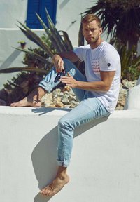 Camiseta blanca con texto rojo, jeans azul claro, descalzo, sentado en una pared blanca, rodeado de vegetación, con gafas de sol en la mano.
