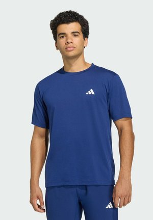 Jeune homme portant un t-shirt et un short Adidas bleus assortis avec des logos Adidas blancs, debout devant un fond clair uni.