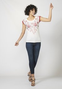 T-shirt blanc avec un motif floral rouge, manches courtes rouges, associé à un jean skinny bleu foncé et des sandales noires à brides.