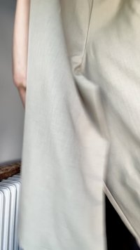 Beige, lightweight fabric draping around a figure, showing soft texture and smooth finish. A wooden surface and radiator are partially visible.