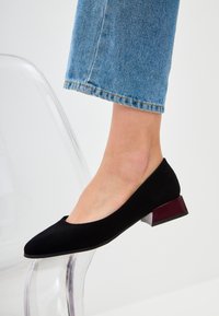 Décolleté in suede nero con punta arrotondata e tacco basso a blocco con design trasparente e accento bordeaux. Indossato con jeans azzurri.
