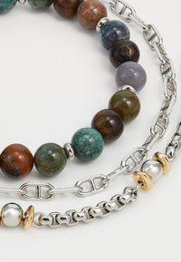 Trois colliers : un avec des perles rondes multicolores, un deuxième en maillons de chaîne en argent, et un troisième avec des accents métalliques argentés et dorés.