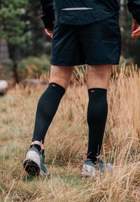 Calze a compressione nere fino al ginocchio indossate con pantaloni corti neri e scarpe da ginnastica grigie, ambientate in uno sfondo naturale e erboso.