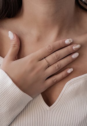 Gold ring with small gemstones on a finger, positioned over a neckline in a ribbed, light-colored top; smooth, well-groomed nails with designs.