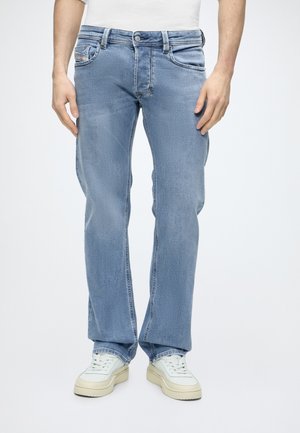 Homme portant un jean droit bleu clair et des baskets blanches, debout les mains détendues le long du corps.