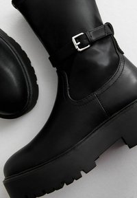 Svarta läderankelboots med en tjock plattformssula, spänndetalj nära toppen och slät textur med synliga sömmar.