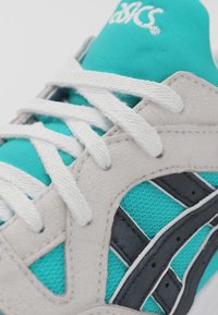 Närbild av en teal och beige ASICS-sko med vita snören och svarta ränder på sidan.