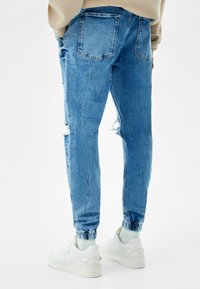 Pantalones jogger de mezclilla azul claro con detalles desgastados, puños elásticos y bolsillos traseros, combinados con zapatillas blancas.