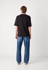 Persona con cabello corto marrón, vestida con una camiseta negra de manga corta simple y jeans azules holgados, de pie mirando hacia atrás sobre un fondo blanco.