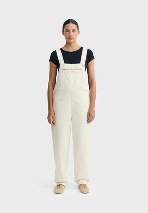 Mujer de pie mirando al frente, vestida con un overol color crema con un bolsillo delantero y una camiseta negra de manga corta debajo, sobre un fondo liso.