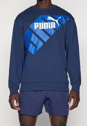 Sudadera de algodón azul marino con un gran diseño del logo de Puma en azul y blanco, cuello redondo y puños y dobladillo acanalados. Llevada con pantalones cortos a juego.
