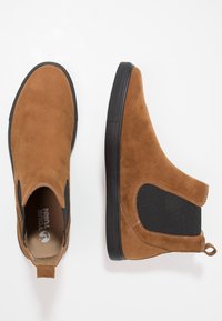 Brązowe zamszowe botki do kostki z elastycznymi panelami bocznymi, teksturowaną gumową podeszwą i małą pętelką z tyłu. Gładkie wykończenie z detalami zszycia.