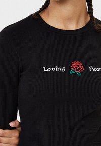Svart ribbad långärmad skjorta med vit broderad text "Loving" och "Heart" samt en röd rosdesign mellan orden.