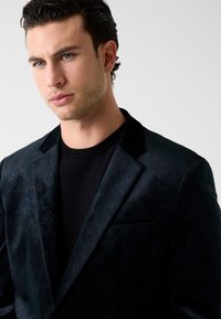 Zwart patroonblazer met een subtiele textuur, voorzien van een enkele zak en een inkeping in het revers, gedragen over een zwart T-shirt met ronde hals.