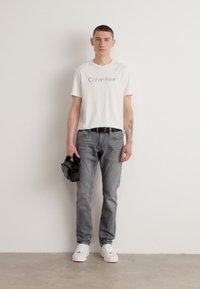 Balta t-kreklu ar "Calvin Klein" logotipu, pelēkās biksēs, baltās sporta kurpēs, tur melnu somu, stāvot pret neitrālu fona.