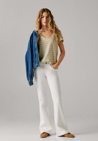 Moteris su ilgais bangeliais, dėvinti dryžuotą smėlio spalvos viršutinį drabužį, baltas išplatėjančias kelnes, rudos spalvos batus ir nešiojanti ant peties mėlyną džinsinę striukę.