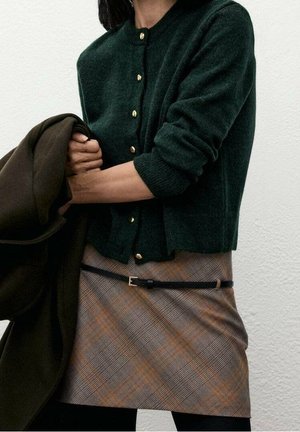 Frau trägt dunkelgrüne, geknöpfte Strickjacke und braunen karierten Rock mit schwarzem Gürtel, hält einen dunkelgrünen Mantel vor hellem Wandhintergrund.