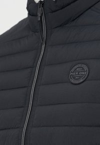 Gros plan d'une veste matelassée noire avec une fermeture éclair frontale et un logo rond portant l'inscription Pier One sur la poitrine.