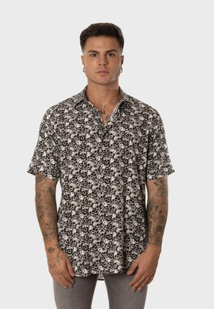 Hombre con el cabello corto y oscuro que lleva puesta una camiseta de manga corta con estampado de calaveras en blanco y negro, mostrando tatuajes en ambos antebrazos.