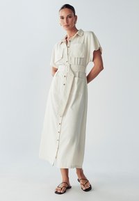 Beige Hemdblusenkleid mit kurzen Ärmeln, Gürtel in der Taille, zwei Vorderaschen und geradem Schnitt, kombiniert mit schwarzen Sandalen.