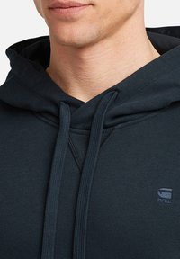 Sudadera con capucha azul marino que presenta un cordón, tejido texturizado y un pequeño detalle de logo en el pecho. Diseño de ajuste clásico.