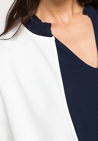 Giacca bianca testurizzata con colletto rotondo, indossata sopra un top blu navy con scollo a V. Cerniera visibile sulla parte anteriore.