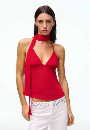 Sötét egyenes hajú nő piros, sálrészletes pántos felsőt és ezüst szegecsekkel díszített fehér nadrágot visel.