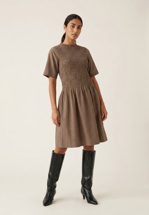 Femme debout portant une robe marron à carreaux longueur genou avec des manches courtes et des bottes noires à talons montant jusqu’aux genoux, sur fond uni.