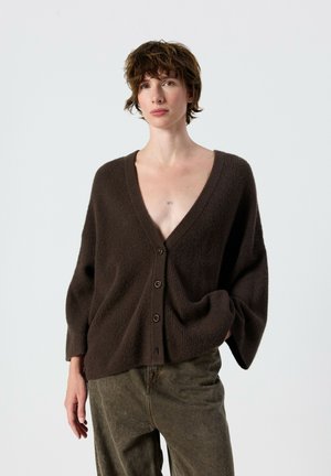 Person with short brown hair wearing a loose dark brown buttoned cardigan and green pants, standing with one hand in pocket.