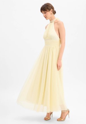 Femme portant une robe maxi jaune pâle à col halter, taille cintrée et jupe légère et fluide, associée à des sandales dorées à talons hauts.