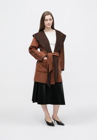 Palton din amestec de lână cu curea, cu guler adânc în V, blocuri de culoare maro și ruginiu, buzunare mari și o broșă decorativă pe rever.