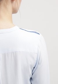 Ljusblå blus, mjukt tyg, knappdetaljer vid axlarna, rund halsringning och sömlös bakdesign som framhäver en j ämn textur.