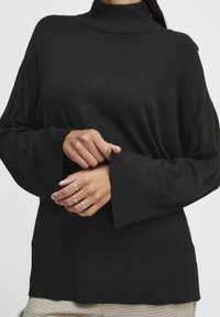 Pulover negru cu guler înalt, cu un croi lejer, mansete cu textură și un corp texturat. Detalii notabile includ o margine lata și un material moale.