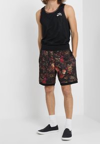Débardeur en mesh noir avec logo blanc. Shorts noirs à motif floral avec bordures noires. Chaussures noires sans lacets et chaussettes blanches à la cheville.