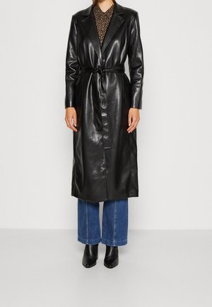 Personne portant un long manteau en cuir noir ceinturé par-dessus un jean bleu et des bottines noires, debout devant un fond uni.