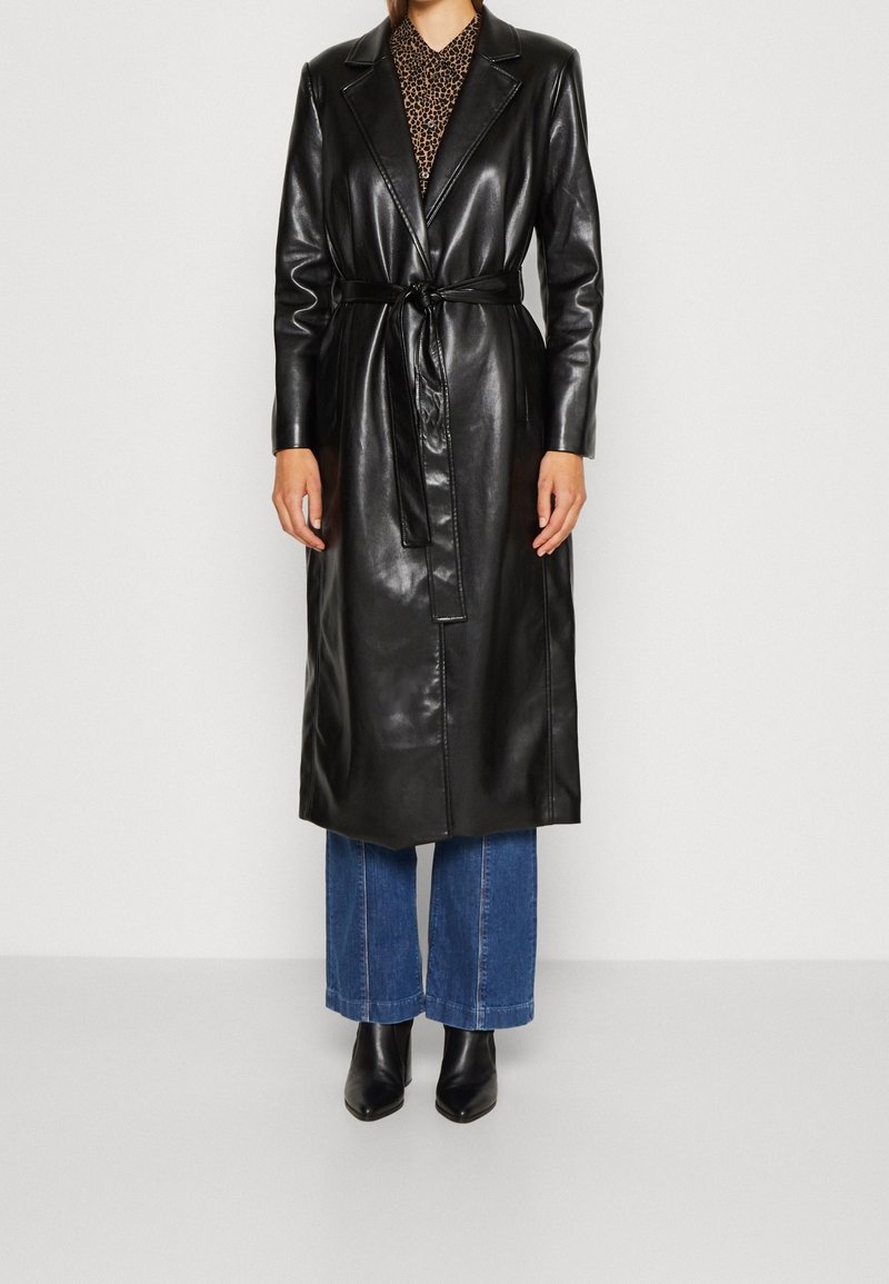 Personne portant un long manteau en cuir noir ceinturé par-dessus un jean bleu et des bottines noires, debout devant un fond uni.