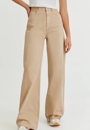 Pantalon beige à jambes larges porté avec des baskets blanches, haut blanc court, une main sur la hanche et l'autre détendue sur le côté.