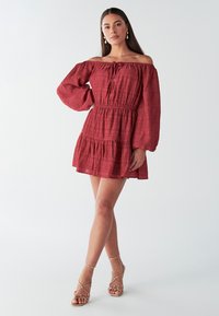 Rode textuur jurk met blote schouders, lange mouwen, elastische tailleband en gelaagde rok. Gecombineerd met gouden sandalen met bandjes.