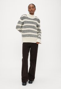 Žena stojící, oblečená v krémovém a černém pruhovaném pleteném svetru, tmavě hnědých širokých kalhotách a černých botách na jednoduchém světlém pozadí.